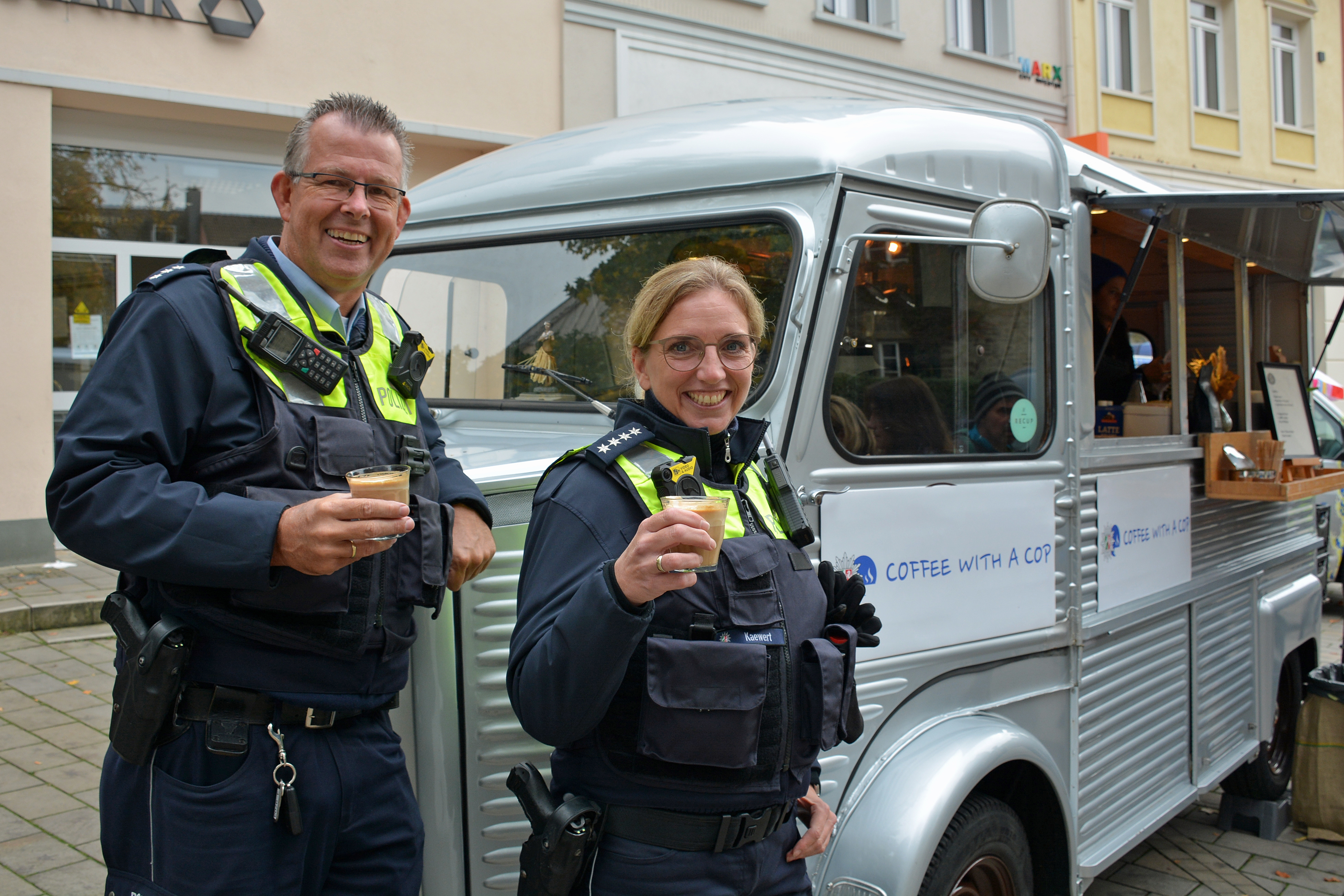 A policeman and a policewoman drink a coffee in front of a historic coffee mobile.