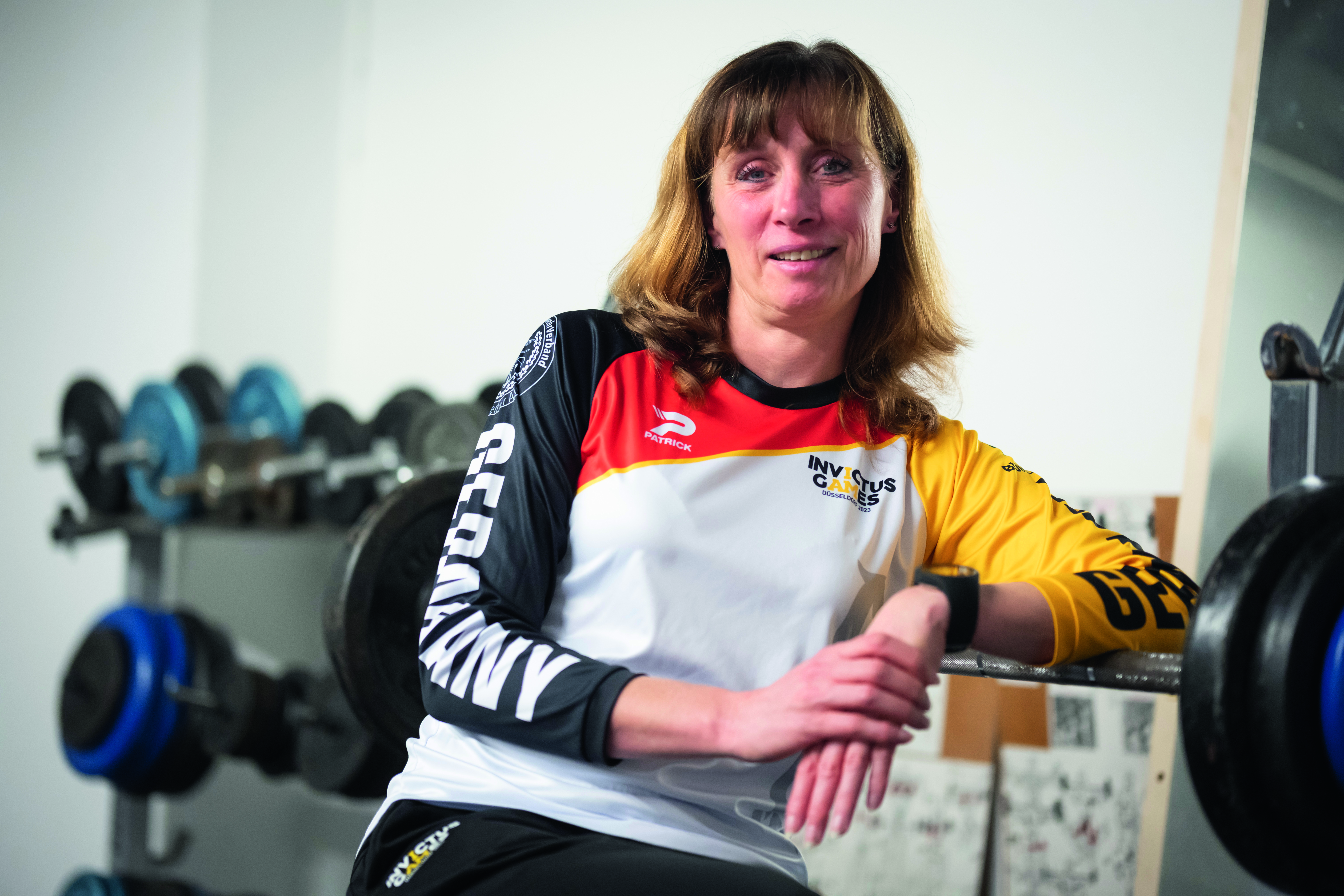 Im Vordergrund sitzt Polizistin Sandra Müller. Sie befindet sich in einem Trainingsraum und trägt ein Trikot der Invictus Games. Im Hintergrund sind Gewichte auf Halterungen zu sehen.